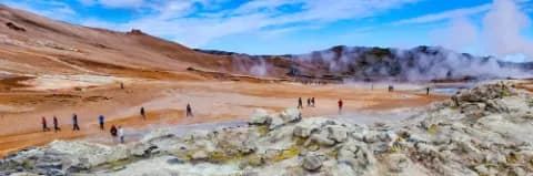 Mehrere Menschen gehen auf einem weiten, dampfenden Geothermalfeld in Island mit Felsen und Hügeln.