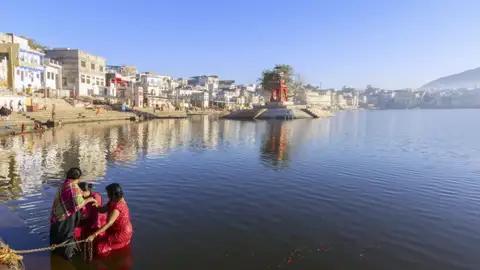 Drei Personen in farbenfrohen Saris bei einer rituellen Waschung im heiligen Pushkar-See, einem Highlight der Rajasthan-Reise, mit den Ghats und der Stadt Pushkar im Hintergrund.